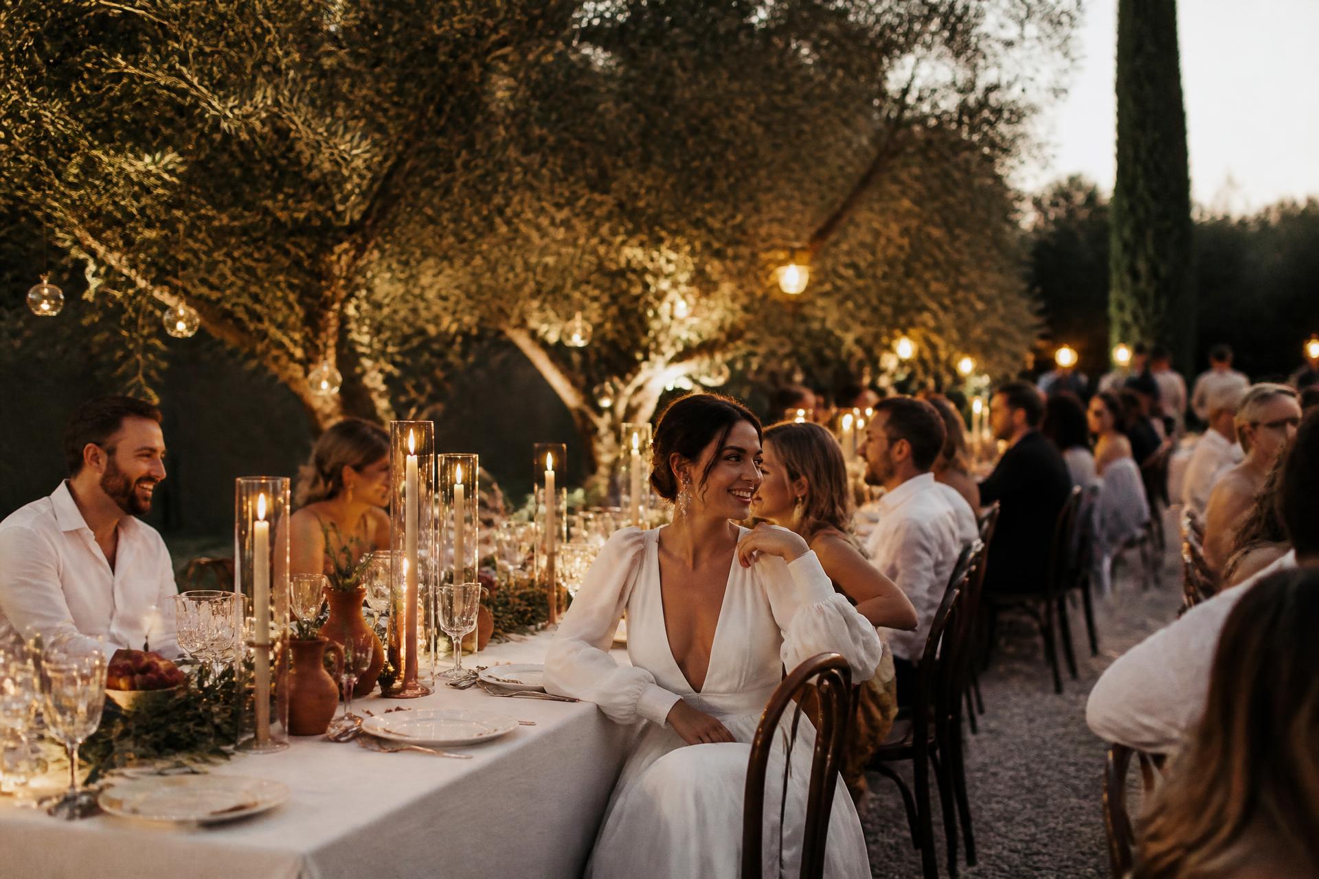 Guests seated at a long candlelit table beneath olive trees at dusk